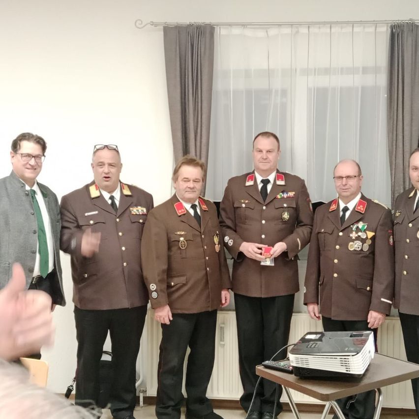 Sechs Männer in Militäruniformen stehen nebeneinander. Sie posieren für ein Foto. Sie stehen vor einer weißen Wand mit einem Vorhang. Auf einem Tisch steht ein Projektor mit einer Fernbedienung.
