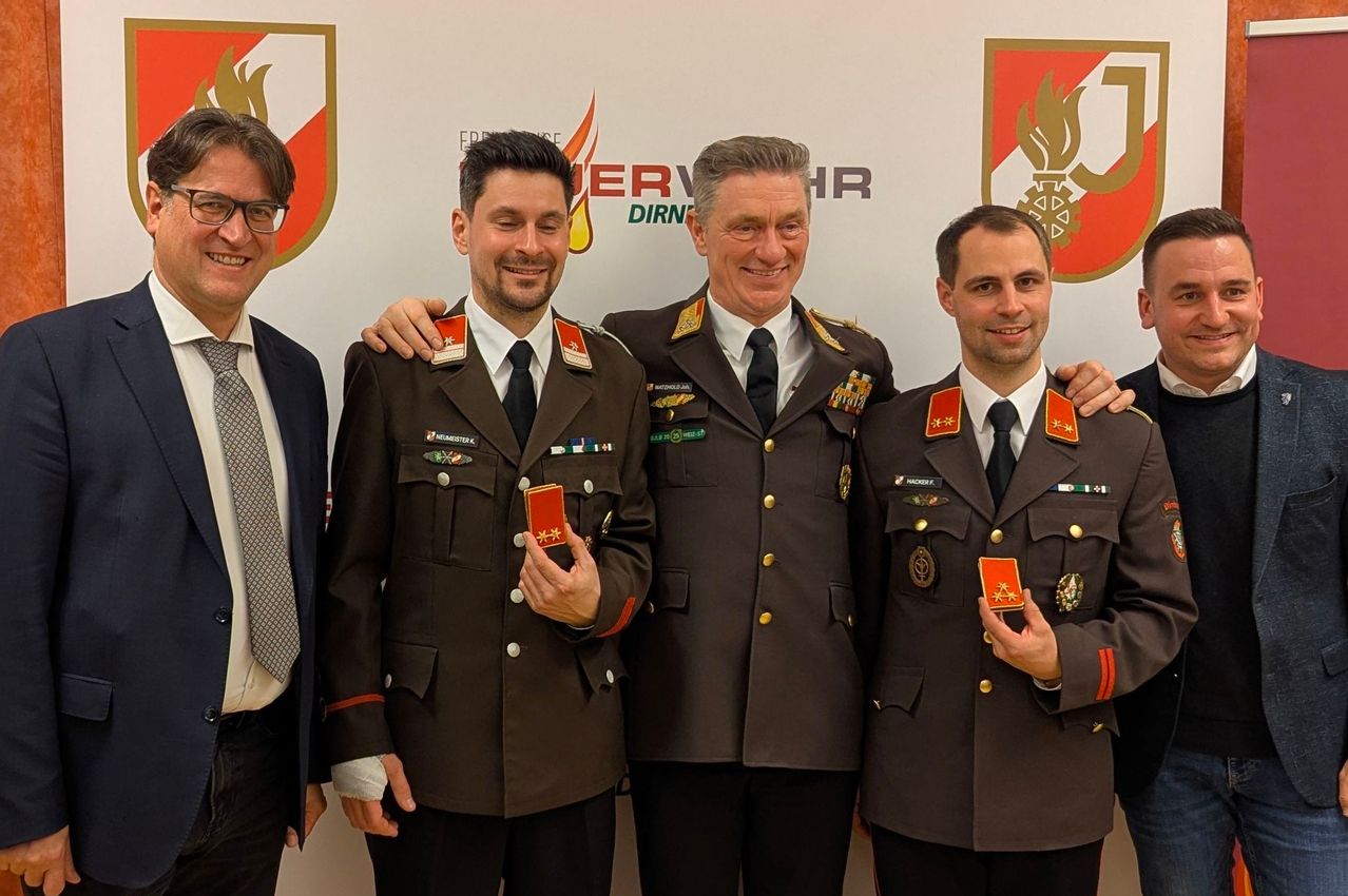 Vier Männer in Militäruniformen posieren für ein Foto. Die beiden Männer in der Mitte haben ihre Arme umeinander gelegt. Sie halten rote Abzeichen in den Händen. Hinter ihnen ist eine weiße Wand mit Logos.