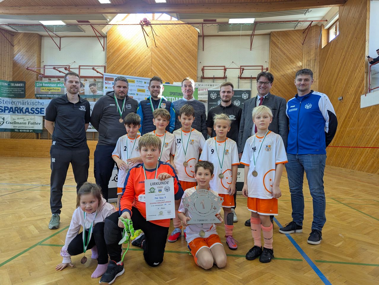 Eine Gruppe von Kindern und Erwachsenen posiert für ein Foto in einer Sporthalle. Die Kinder tragen Trikots mit Nummern und Medaillen. Ein Junge hält ein Zertifikat und einen Pokal. Erwachsene stehen hinter den Kindern. Es gibt Poster an der Wand.