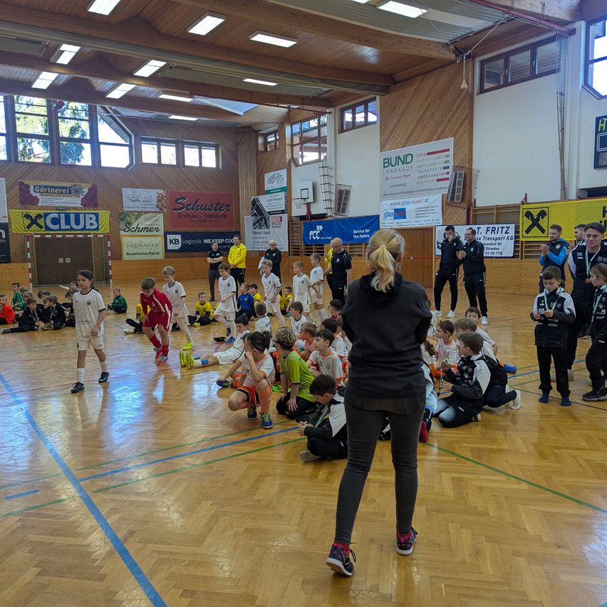 In einer Turnhalle versammeln sich eine Gruppe von Kindern um eine Frau, wahrscheinlich ihre Trainerin, die sie anweist. Einige Kinder stehen, während andere auf dem Boden sitzen. Die Turnhalle hat Holzböden und Banner an den Wänden.