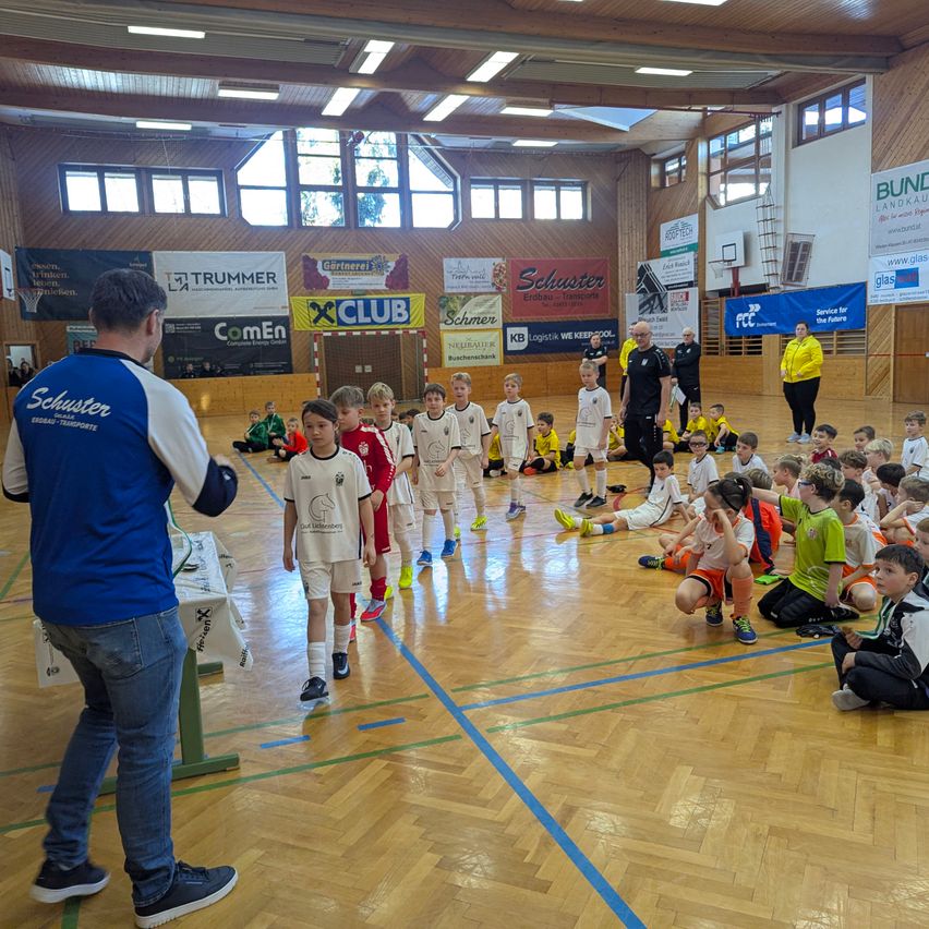 Ein Fußballtrainer spricht mit jungen Spielern auf einem Platz, Zuschauer sitzen auf dem Boden und Banner hängen an der Wand.