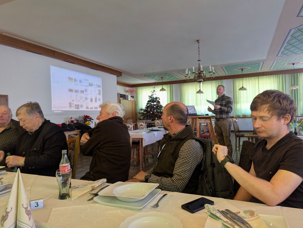 In einem Esszimmer sitzen Menschen um einen Tisch herum. Ein Mann steht vor einer Projektionsleinwand und hält eine Präsentation. Ein anderer Mann schaut sich die Präsentation an, während ein anderer Mann sein Smartphone betrachtet.