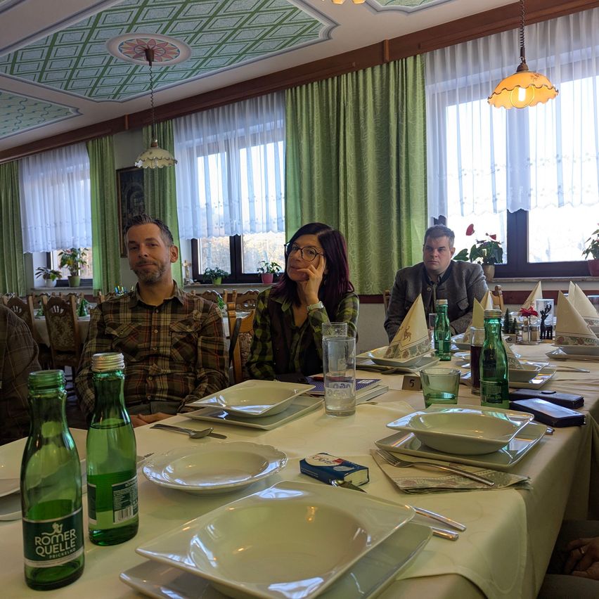 Vier Personen sitzen an einem langen Tisch in einem Restaurant, mit zwei Flaschen Wasser und einer Schachtel Zigaretten. Der Tisch ist mit Tellern, Gläsern und Besteck gedeckt. Hinter ihnen sind grüne Vorhänge und Fenster.