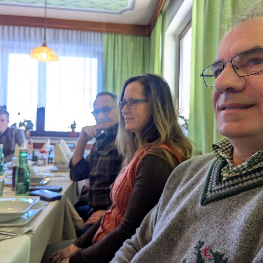 Drei Menschen sitzen an einem Tisch in einem Restaurant. Zwei Männer und eine Frau. Die Frau trägt eine Brille und einen Schal. Der Mann neben ihr trägt eine Brille und ein kariertes Hemd. Der Mann auf der rechten Seite trägt eine Brille und einen Pullover mit grünem Muster.