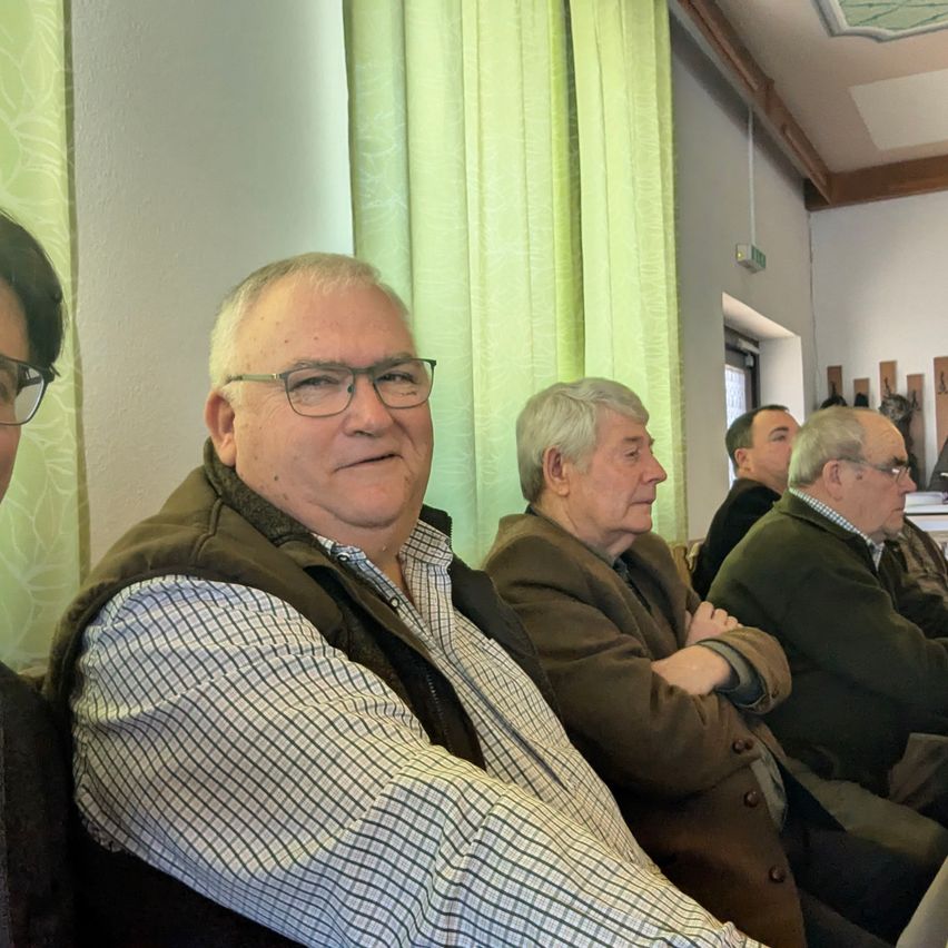 Mehrere ältere Männer sitzen in einem Raum mit grünen Vorhängen. Ein Mann trägt eine Brille und eine braune Weste.