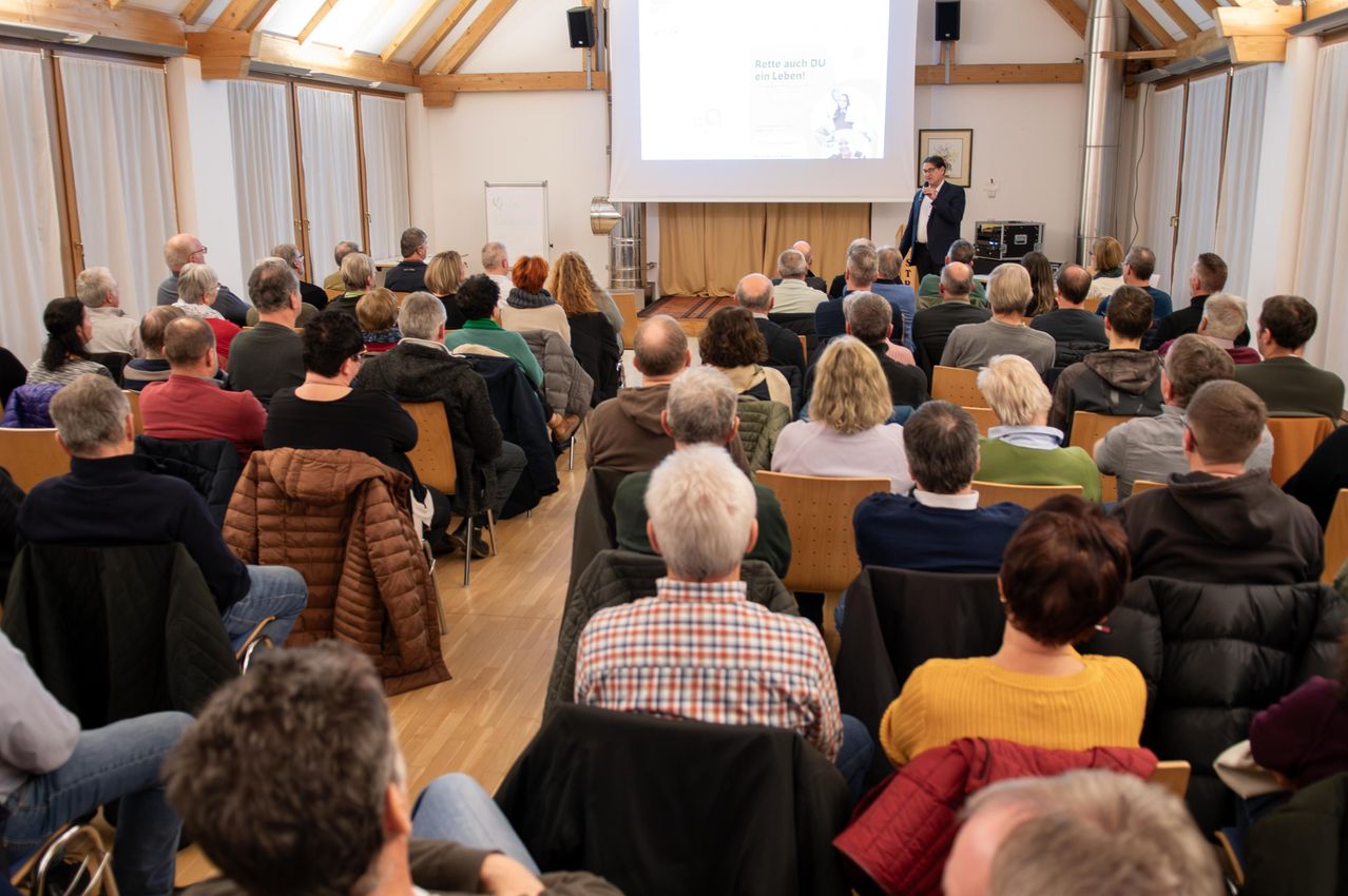 Ein Mann präsentiert vor einem sitzenden Publikum in einem Konferenzraum. Eine Leinwand zeigt eine Folie hinter ihm an.