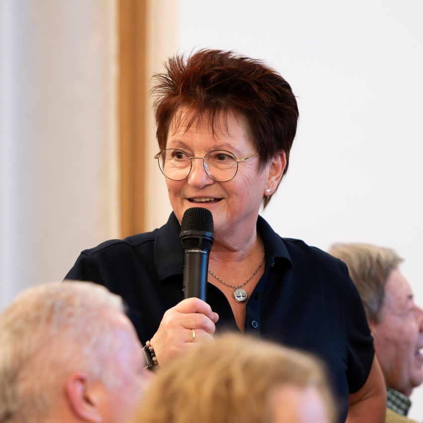 Eine Frau mit kurzen Haaren und einer Brille spricht in ein Mikrofon bei einer Versammlung. Sie lächelt und trägt eine Halskette und ein blaues Hemd. Ein Mann und eine weitere Person hören hinter ihr zu.