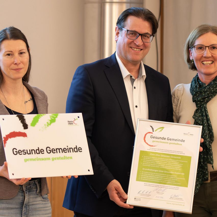 Drei Menschen posieren für ein Foto. Der Mann in der Mitte lächelt und hält ein Zertifikat. Die beiden Frauen auf beiden Seiten halten Schilder. Die Frau auf der linken Seite hält ein großes Schild mit der Aufschrift 'Gesunde Gemeinde'. Der Mann und die Frau auf der rechten Seite halten kleinere Schilder.