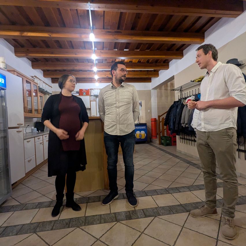 Drei Personen stehen in einem Raum mit gefliesten Böden, Holzbalken und Schränken. Ein Mann hält ein Glas Bier, während zwei andere ihn betrachten.