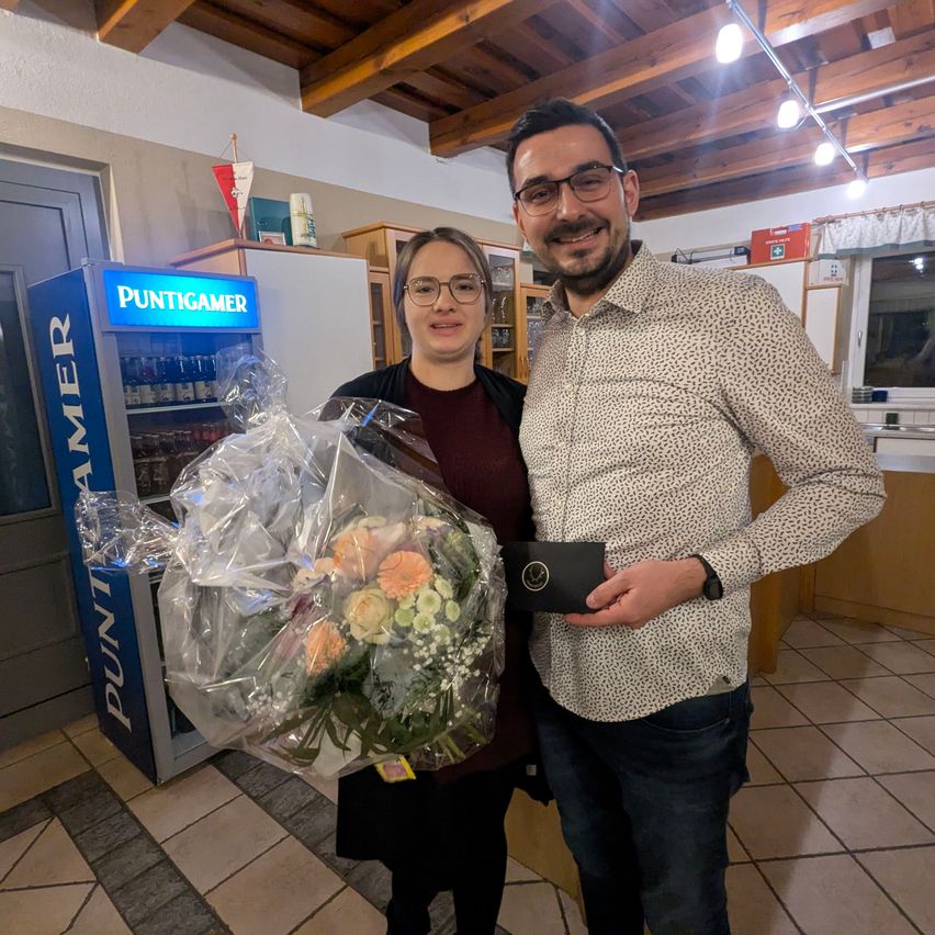 Ein Mann und eine Frau stehen lächelnd nebeneinander, ein Blumenstrauß zwischen ihnen. Hinter ihnen ist ein Verkaufsautomat mit der Aufschrift 'Puntigamer' und ein Kühlschrank zu sehen.