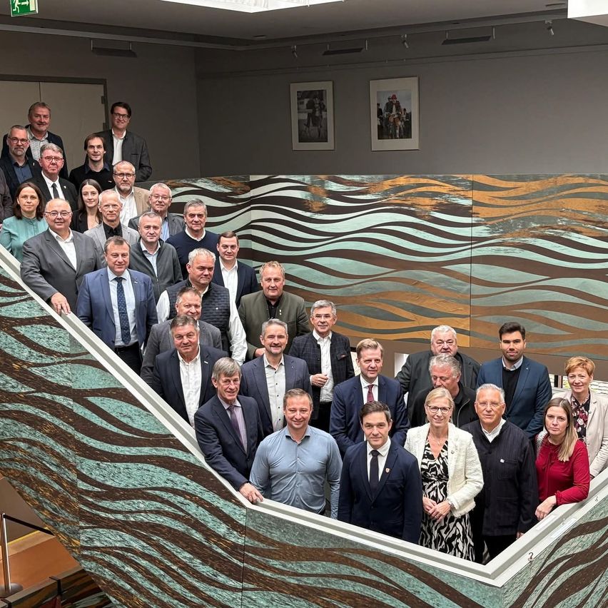Eine Gruppe formell gekleideter Personen steht auf einer Treppe in einem Innenbereich und posiert für ein Foto. Hinter ihnen befindet sich eine Wand mit zwei gerahmten Bildern. Die Treppe hat eine dekorative Wand mit wellenförmigen Mustern.
