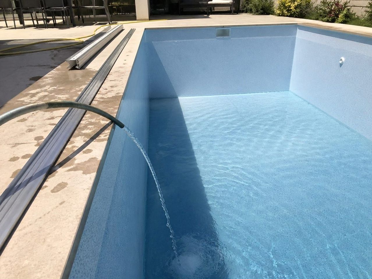 An empty swimming pool with clean blue water is being filled with water through a hose.