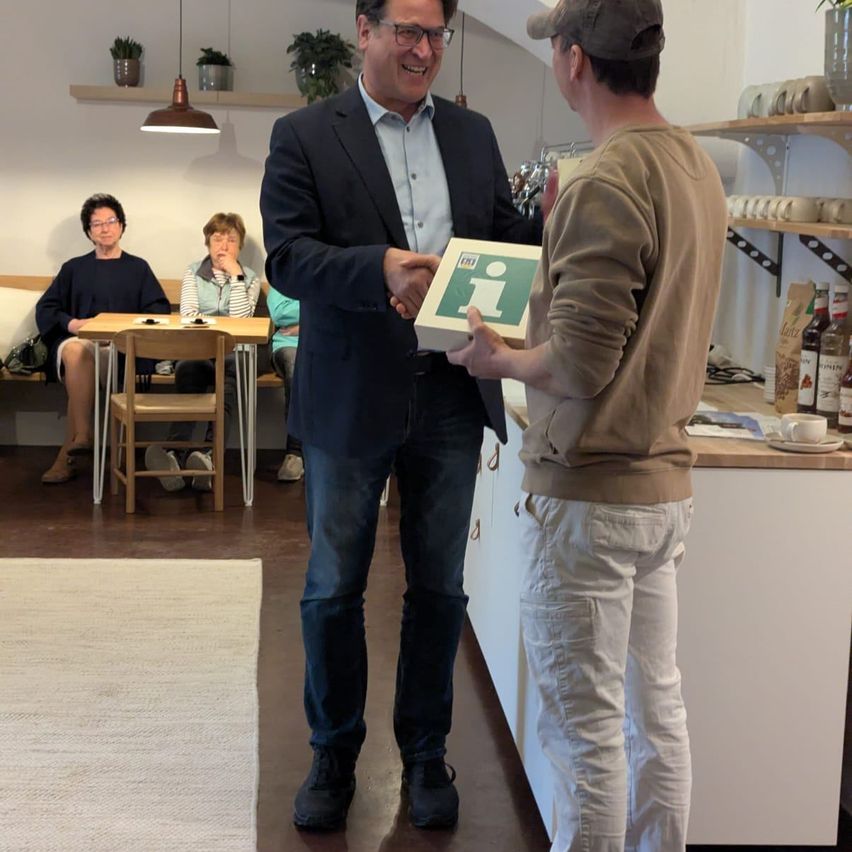 Two men exchange a box in a cafe. One is smiling, holding the box. Another holds the box's lid. People sit behind them at a table. Shelves and bottles are in the background.