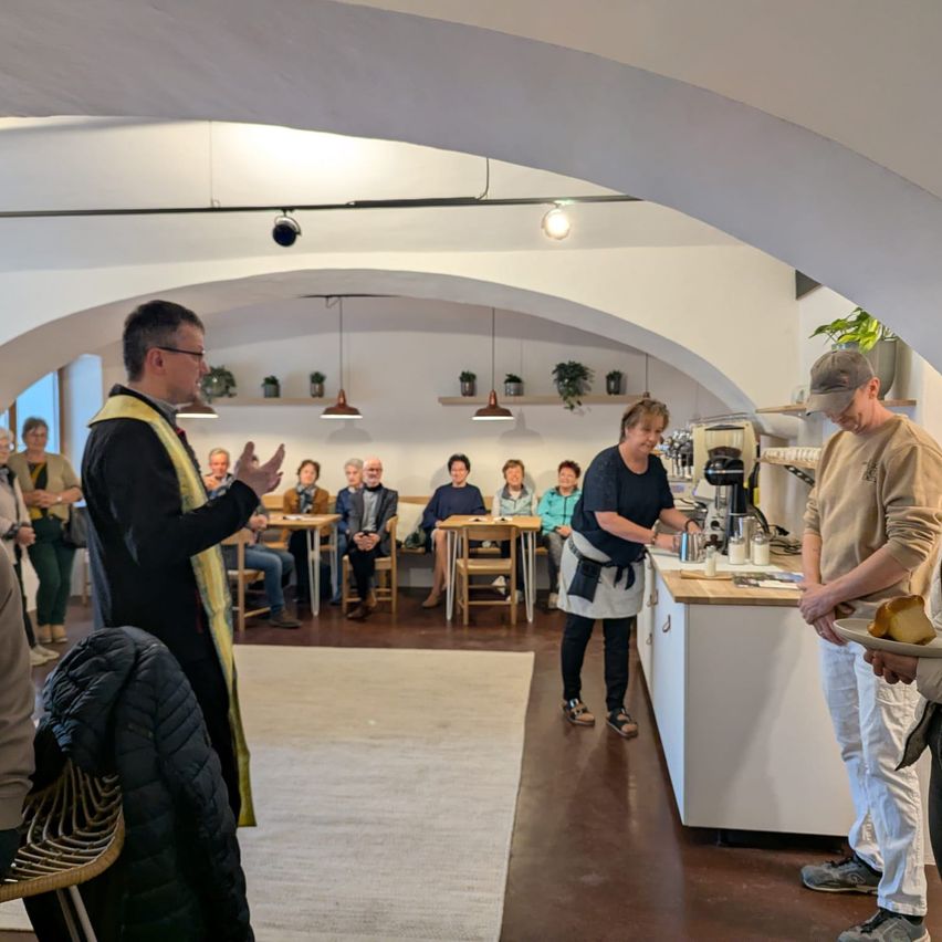 A man in a black robe is standing and talking to a group of people sitting around tables in a room with arched ceilings. A woman prepares coffee at a counter.