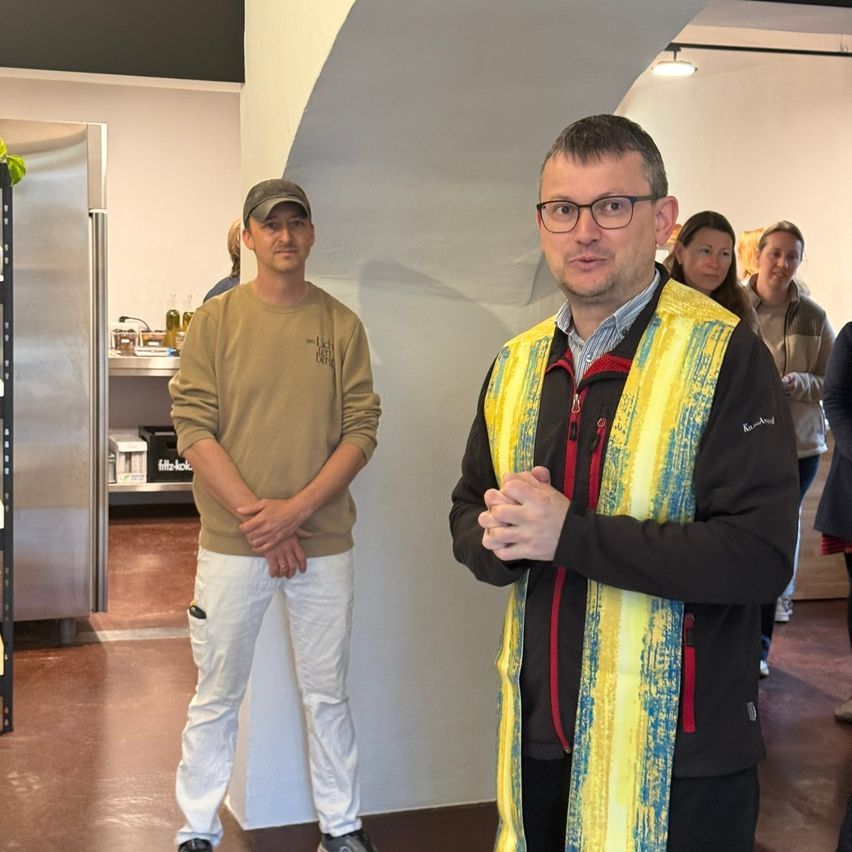 Two men stand in a room, one wearing a reflective vest and the other with a cap. Behind them are shelves with items, and a woman walks nearby.