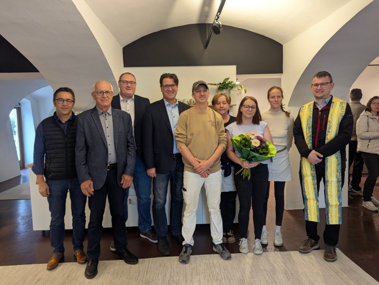 A group of people stands together in a room, smiling for a photograph. One woman holds a bouquet of flowers. The room has white walls and arched ceilings.