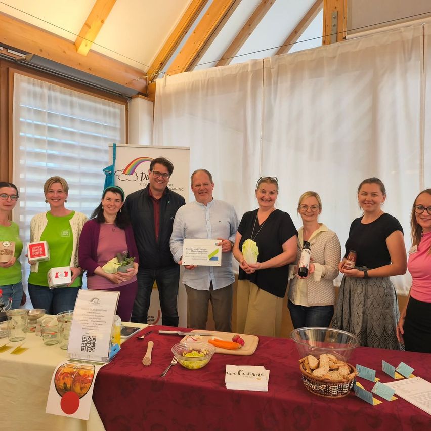Eine Gruppe von Menschen steht um einen Tisch mit Essen und Getränken und lächelt. Ein Mann in der Mitte hält ein Zertifikat. Ein anderer Mann neben ihm hält ein Gemüse. Der Tisch hat Brot, Gemüse und eine Schüssel. Ein Banner mit Text ist im Hintergrund.