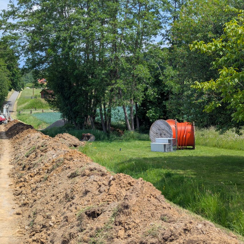 Ein unbefestigter Weg ist von üppigem grünem Laub und Gras umgeben. Ein rotes Baufahrzeug ist auf der linken Seite geparkt. In der Ferne sind ein Haus mit einem roten Dach und ein Feld zu sehen. Ein Bündel orangefarbener Kabel liegt auf dem Rasen.