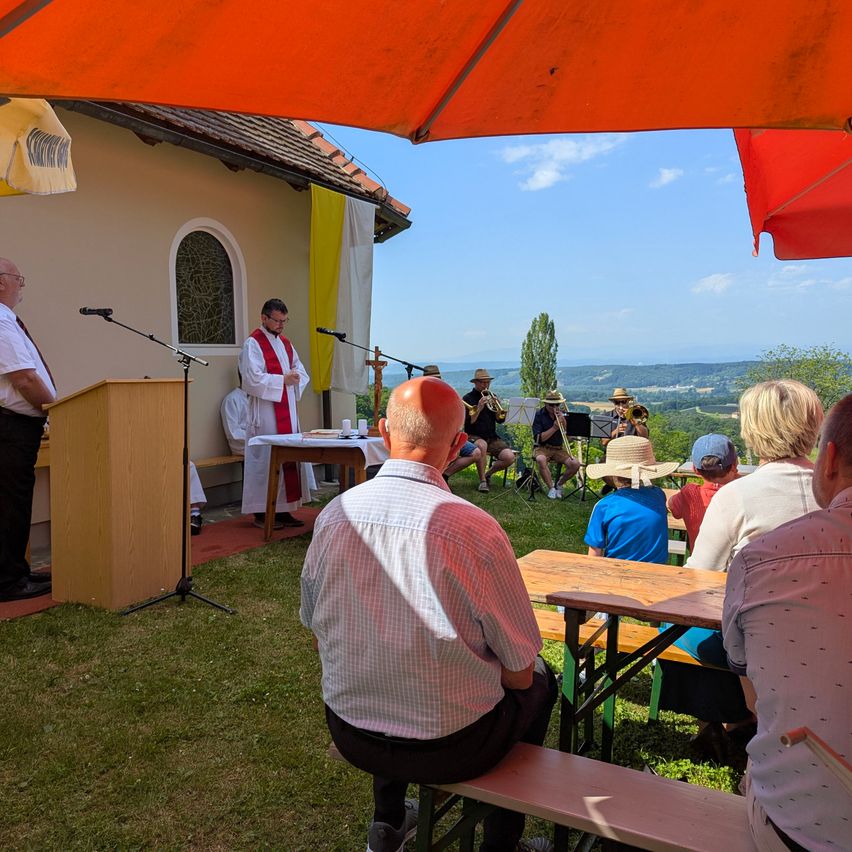 Eine religiöse Zeremonie im Freien. Ein Priester steht an einem Rednerpult mit Mikrofon. Einige Leute sitzen auf Bänken unter einem roten Schirm. Einige Leute sitzen auf dem Gras.