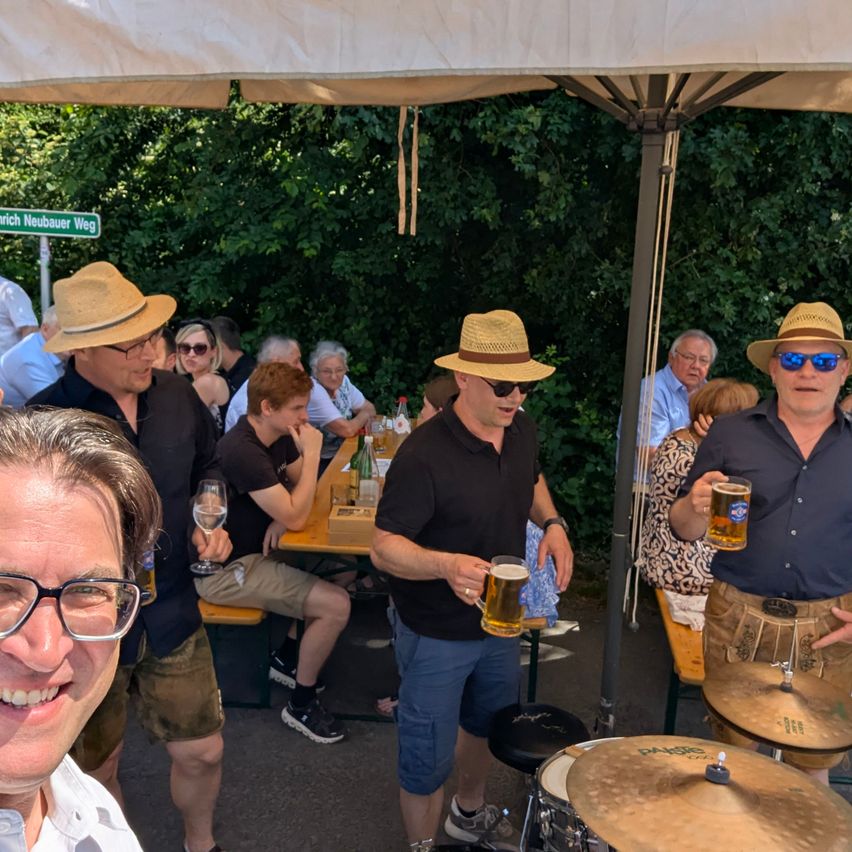 Eine Gruppe von Männern und Frauen genießt Getränke und Essen bei einem Picknick. Ein Mann mit Hut hält ein Bier, während ein anderer mit Brille in die Kamera lächelt.