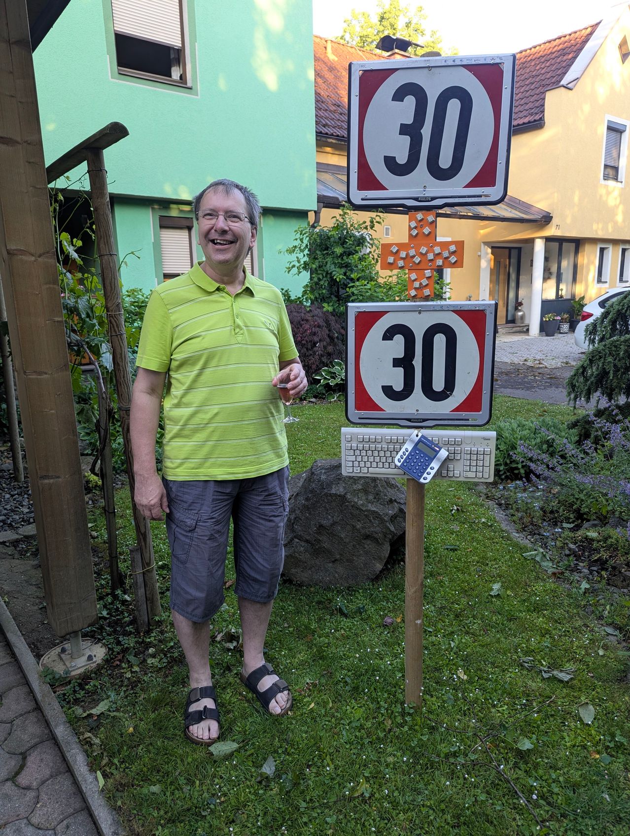 A man in a green polo shirt is smiling in a yard with a speed limit sign.