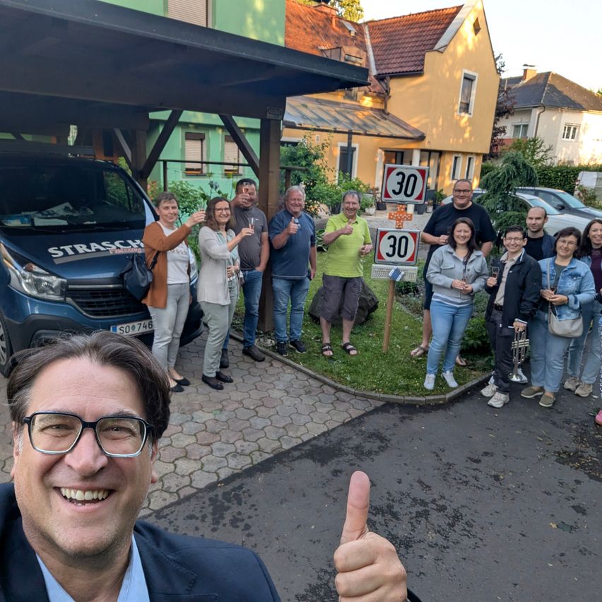 A group of people is gathered in front of a blue van, with some holding glasses. Behind them, a speed limit sign is visible. They are smiling and seem to be posing for a photo.