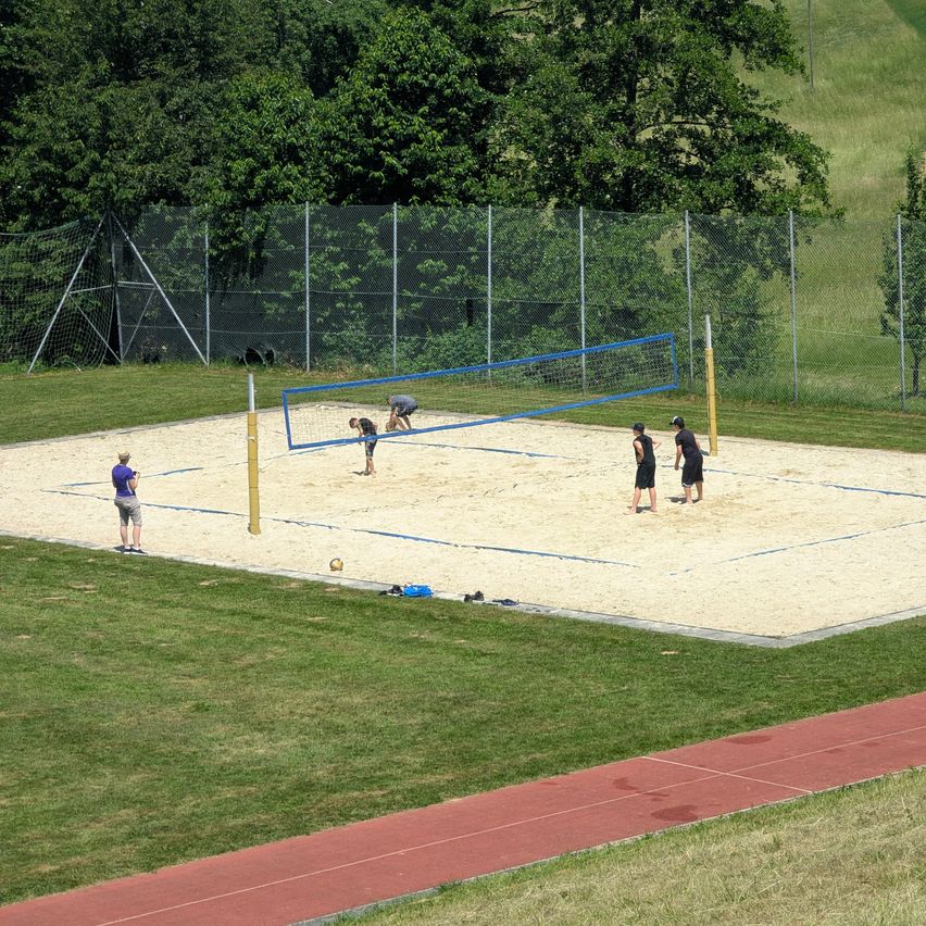 A group of people plays beach volleyball on a sandy court surrounded by a chain-link fence and grass. The players are wearing shorts and sneakers.
