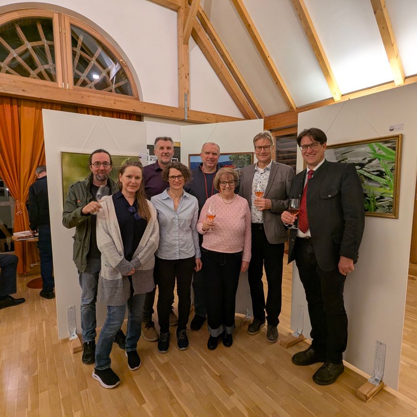 A group of people stand indoors near art displays, posing for a photo while holding wine glasses. The setting features wooden walls and flooring.