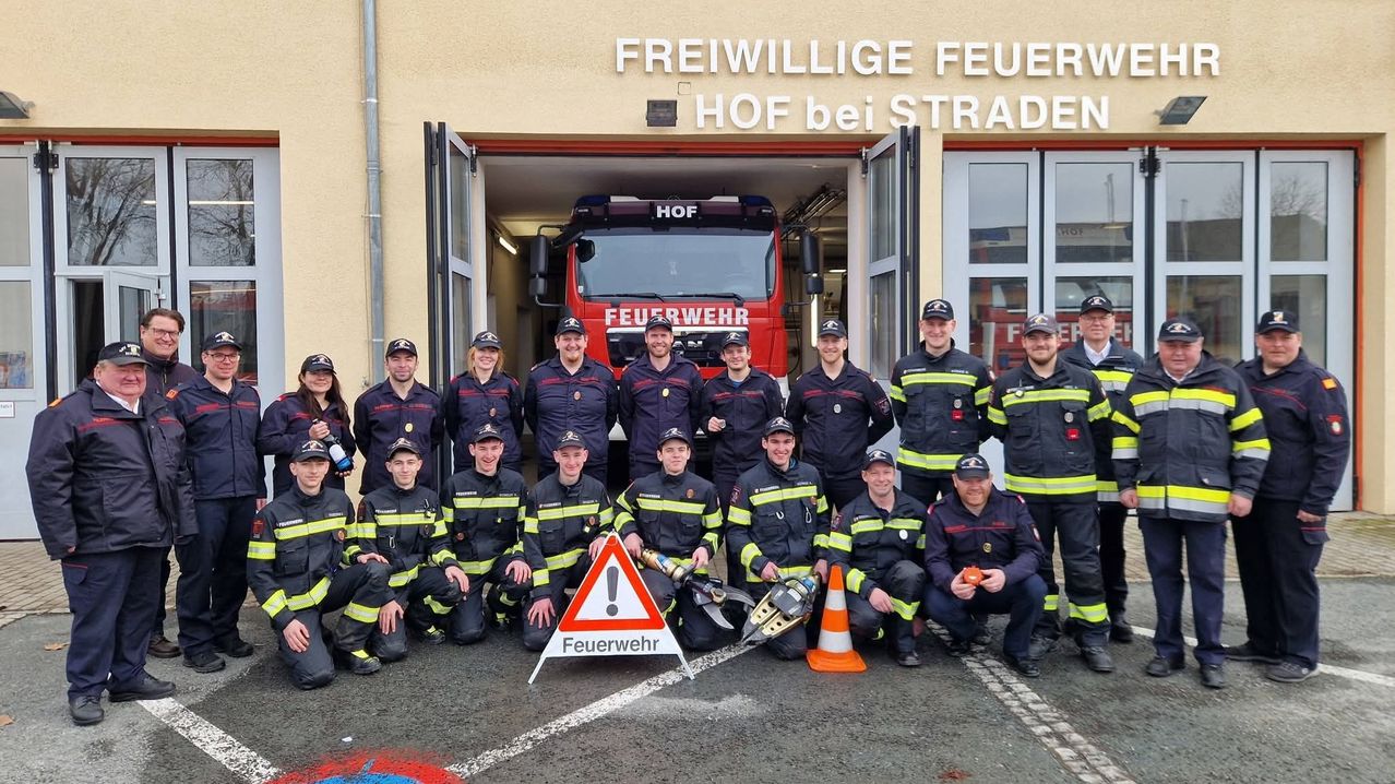 A group of firefighters in uniform pose in front of a fire station with a truck. They are standing in front of a building with open doors.