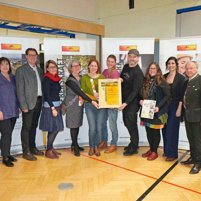A group of people stands on a wooden floor, possibly at an award ceremony. They are holding a certificate, a book, and a cup. Behind them, there are banners with text and images.