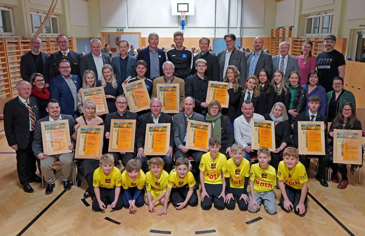 A group of people posing for a photo in a gymnasium, holding awards and certificates, some standing and others sitting on the floor. The background features a basketball hoop.