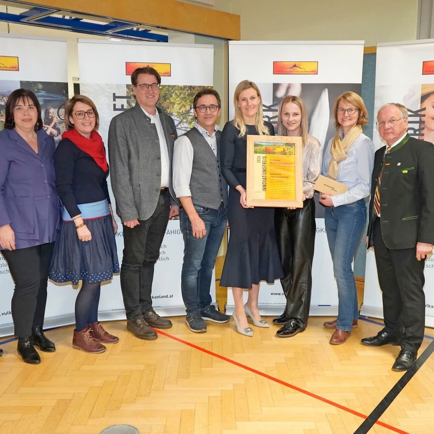 A group of people stand indoors posing for a photo. A woman holds a framed certificate, and another holds a certificate. Two men stand on either side, and three women stand in front of them. Behind them are banners. The floor has a red line.