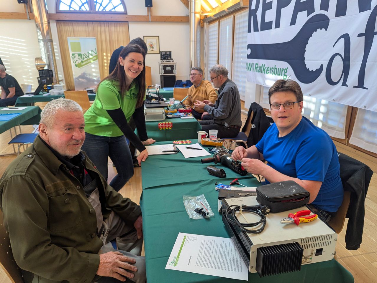 A group of people sit at a table with electronic equipment. A woman stands behind the table, smiling. A man in a blue shirt is working on a device. Another man in a green shirt sits nearby.