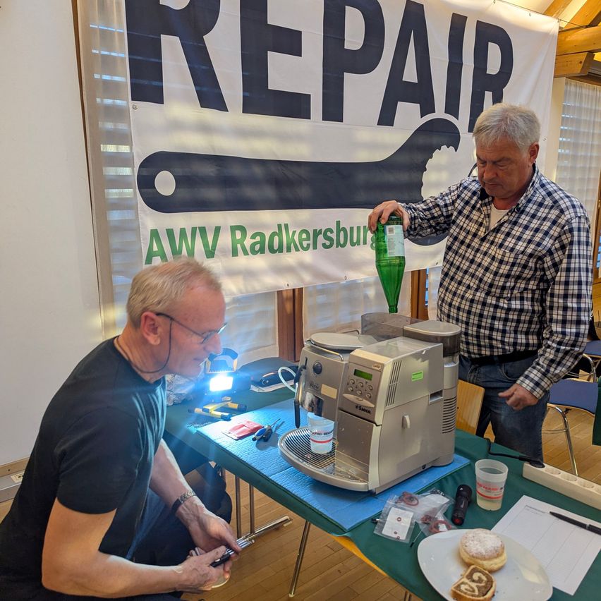 Two men are in a room fixing a machine. One man holds a green bottle while the other examines a device. A banner with the text REPAIR and a wrench logo is visible. On the table, there is a coffee machine, a plate with doughnuts, and other items.