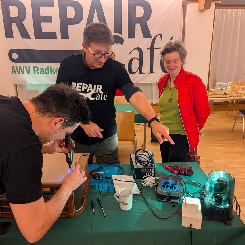 Three people stand in a workshop, with one man using a tool on a radio. Another man points to something while a woman smiles. A banner reads Repair Café.