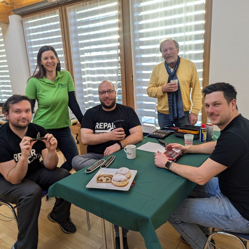 Five individuals are gathered around a green table in a room with wooden and glass doors. Two men are seated, holding tools and a donut plate. A woman stands behind them, and another man stands in the background, all smiling.