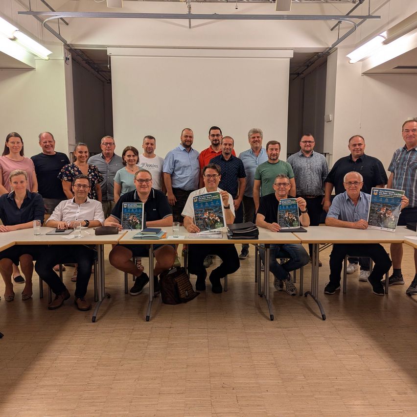 Eine Gruppe von Menschen ist in einem Raum versammelt, sitzt um einen Tisch herum und hält Bücher und Papiere in den Händen. Sie alle lächeln und scheinen für ein Foto zu posieren.