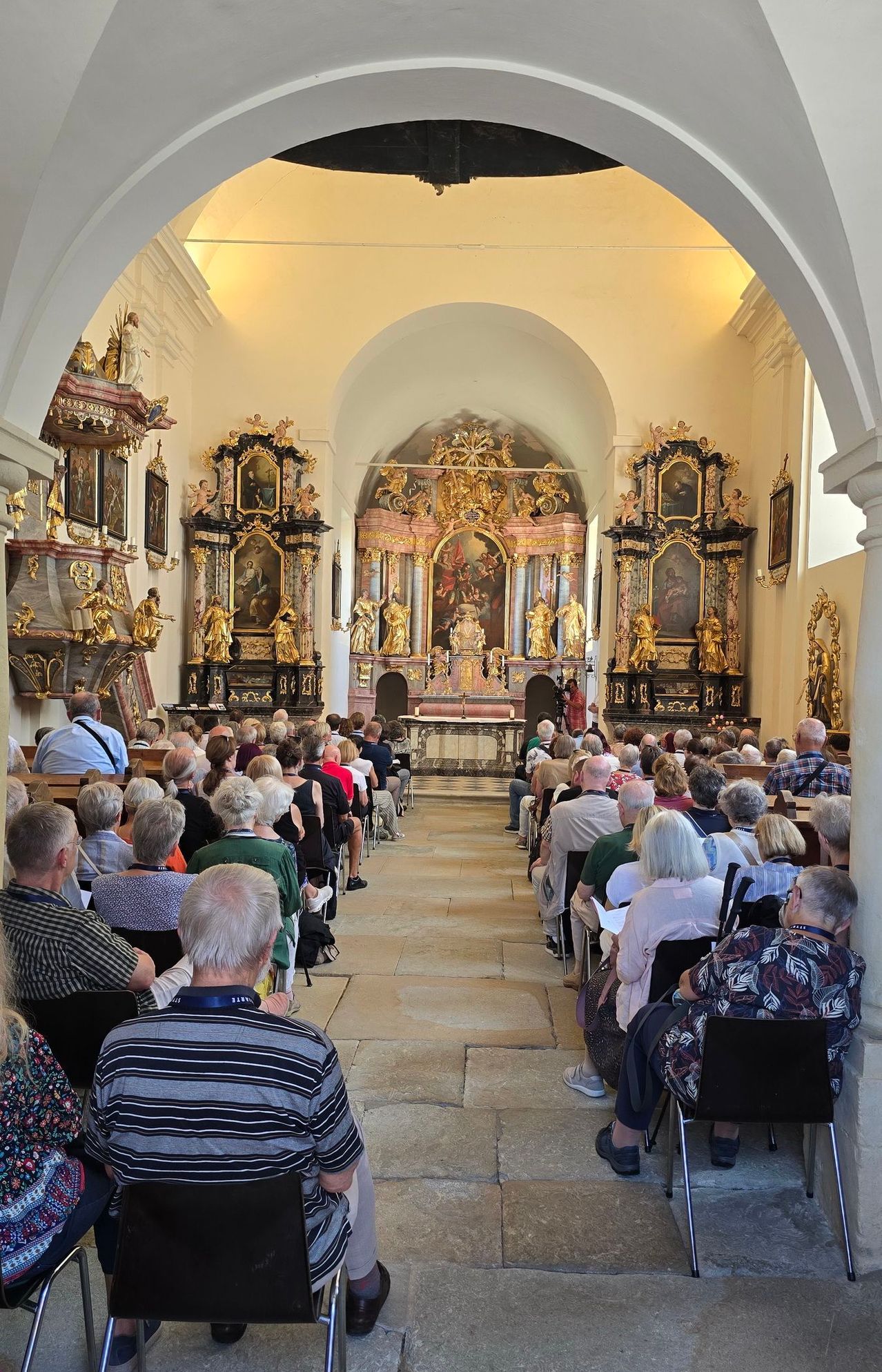 Eine Gruppe von Menschen sitzt in einer Kirche und schaut auf den mit religiösen Kunstwerken und Statuen geschmückten Altar. Das Kircheninnere ist mit aufwendigen Dekorationen und einer hohen Decke ausgestattet.