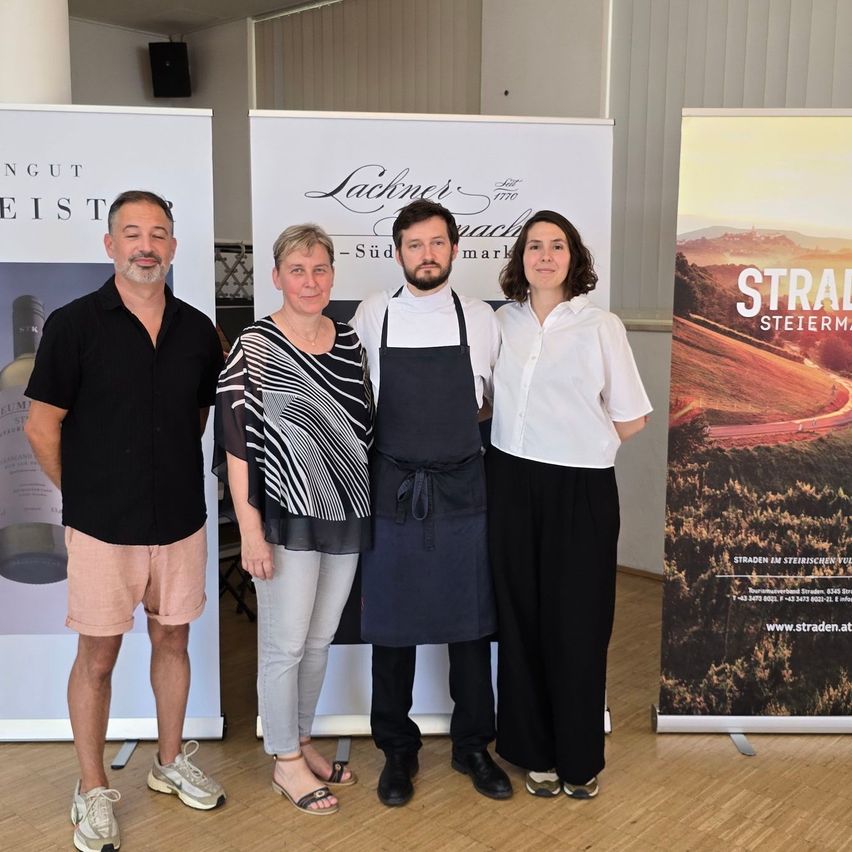 Vier Personen stehen vor einem Hintergrund mit einer Weinwerbung und lächeln für ein Foto. Der Mann links trägt ein schwarzes Shirt und Shorts, während die anderen lässig gekleidet sind. Dahinter zeigt ein Banner eine ländliche Landschaft.
