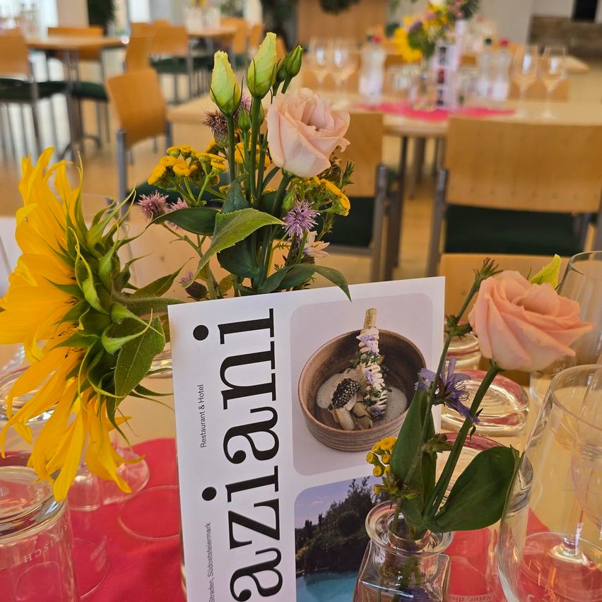 Ein Tisch mit einem Menükarten, Blumen und Weingläsern in einem Restaurant. Die Menükarte lautet 'Tunazz'.