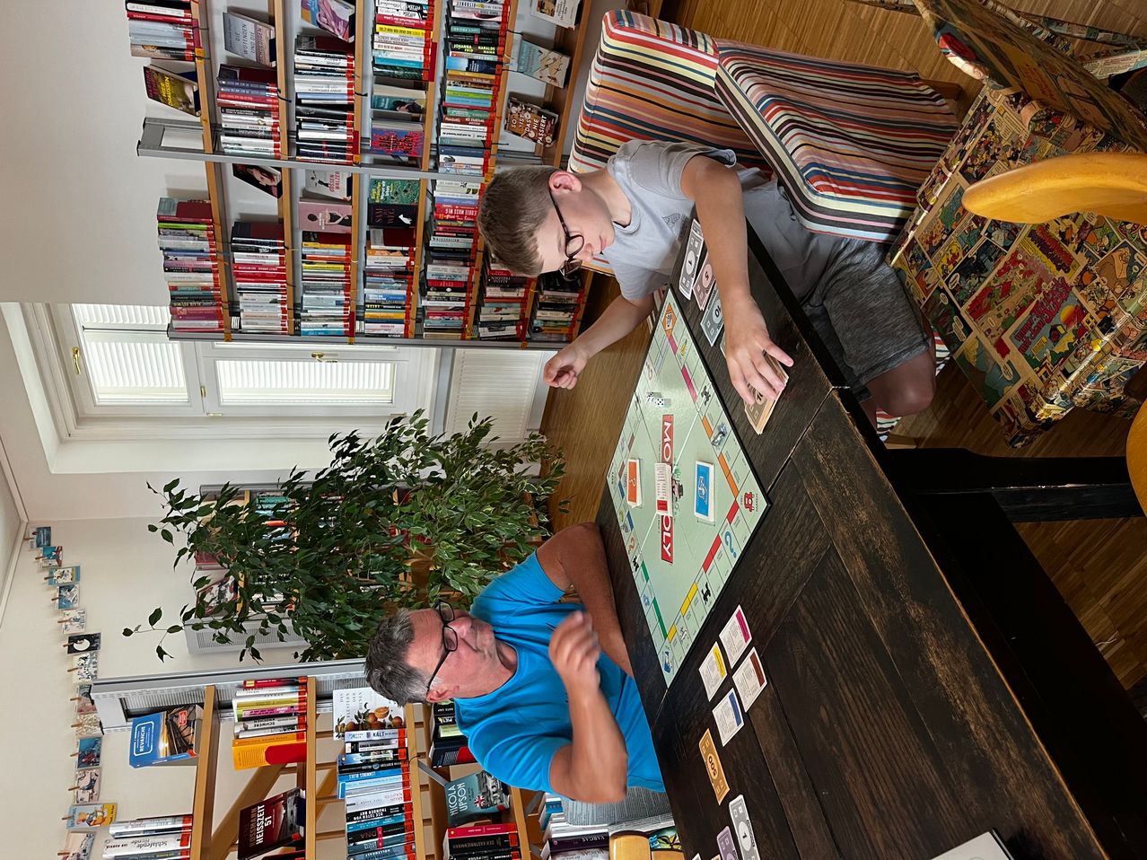 Zwei Männer spielen ein Brettspiel auf einem Holztisch in einem mit Büchern und einer Pflanze gefüllten Raum.