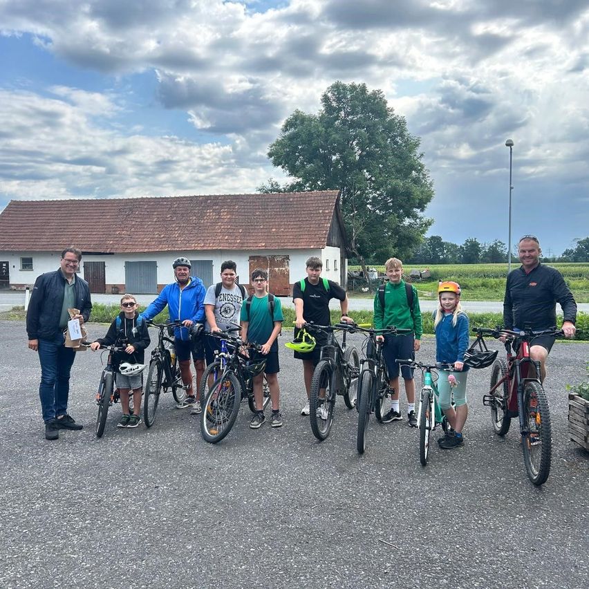 Eine Gruppe von Radfahrern, einschließlich Kindern, posiert für ein Foto vor einem rustikalen Gebäude unter einem teilweise bewölkten Himmel.