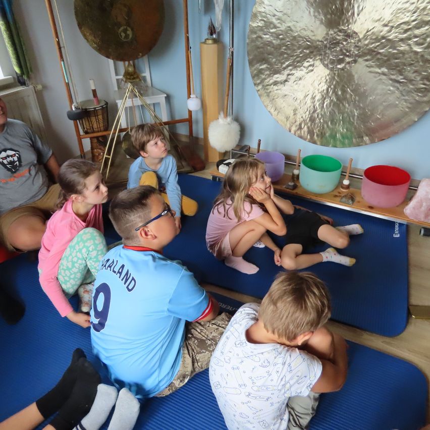 Eine Gruppe von Kindern sitzt auf blauen Matten in einem Raum, einige tragen Socken und Brillen. Ein großer Gong hängt an der Wand.