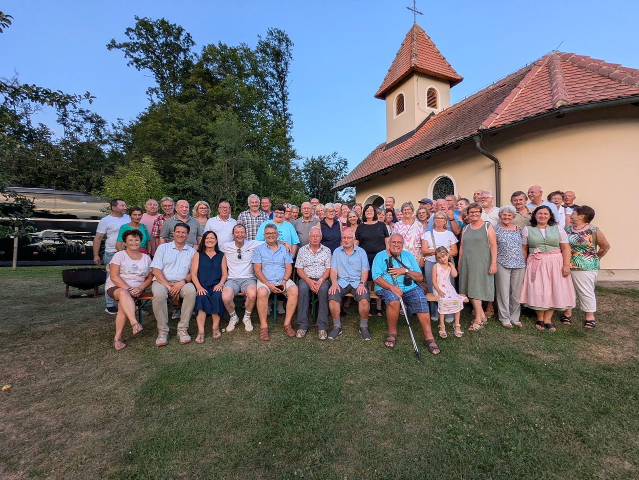 Eine Gruppe von Menschen versammelt sich vor einer Kirche und posiert für ein Foto. Die Kirche hat einen Turm und ein rotes Dach. Bäume sind im Hintergrund sichtbar.