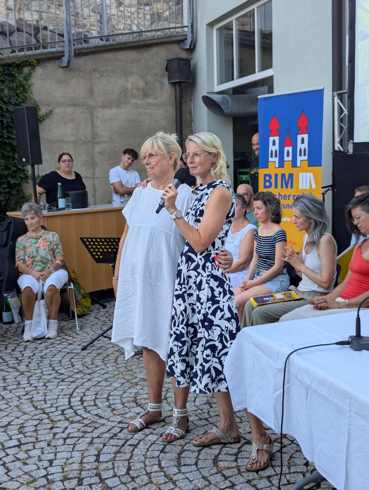Zwei Frauen stehen auf einer Bühne, eine hält ein Mikrofon, während andere in Stühlen hinter ihnen sitzen. Ein Banner mit dem Text 'BIM' ist im Hintergrund sichtbar.