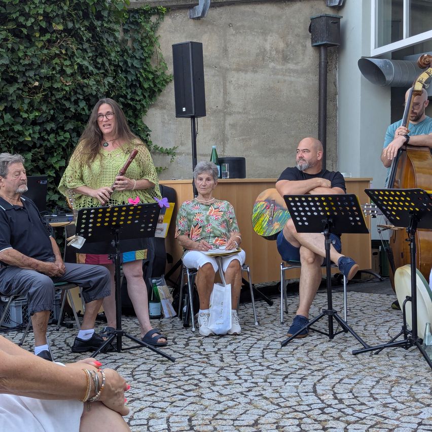 Eine Gruppe von Musikern versammelt sich draußen, eine Frau spielt Flöte und andere spielen verschiedene Instrumente. Sie sitzen um einen Tisch mit Notenständern.