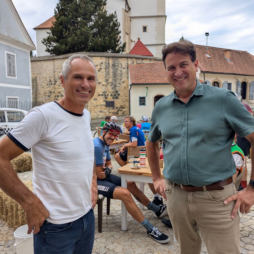 Two men stand smiling in front of a medieval town, with a cyclist resting in the background.