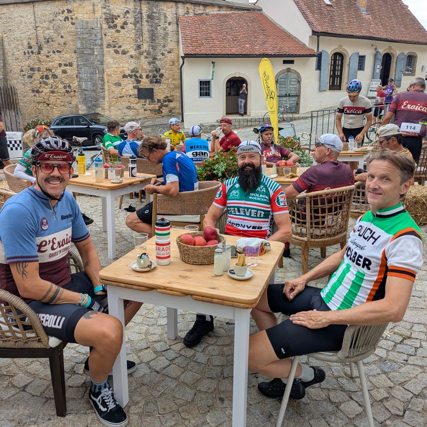 Three cyclists in cycling jerseys sit at a table outside, smiling and posing for a photo. They are surrounded by other cyclists and a building with a red roof.