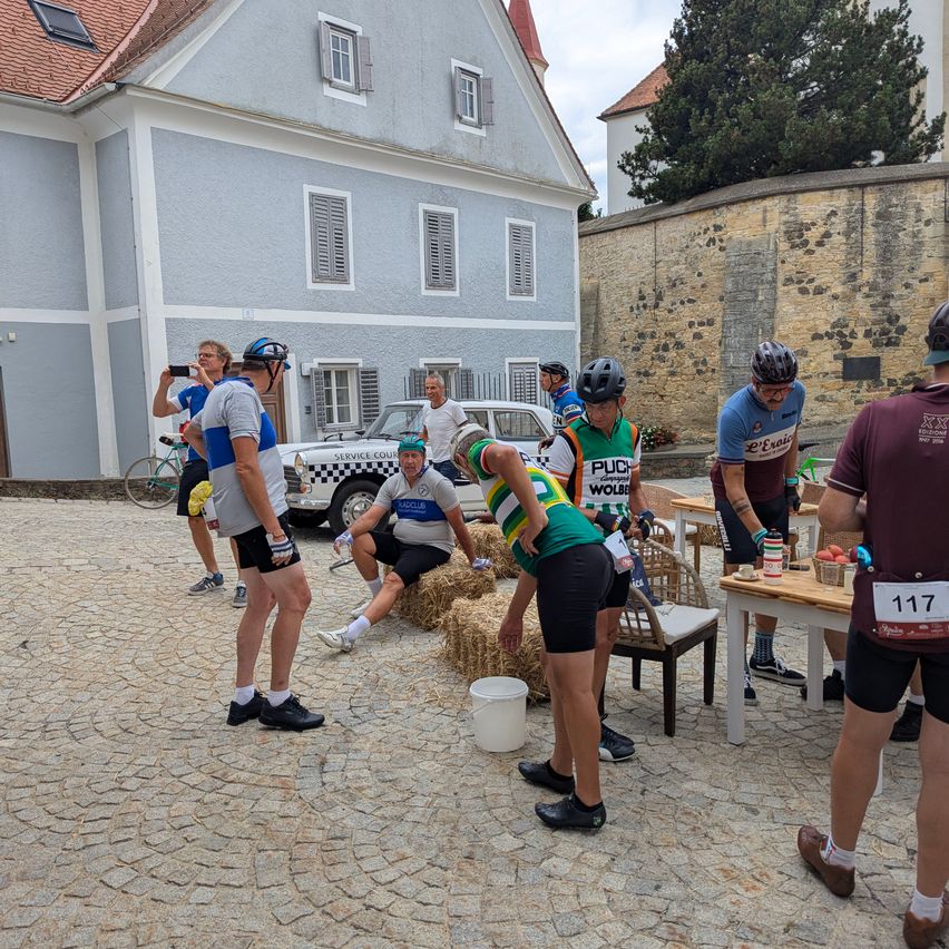 A group of cyclists in cycling gear are gathered around a table with food and drinks. They are in front of a building with a car parked nearby.