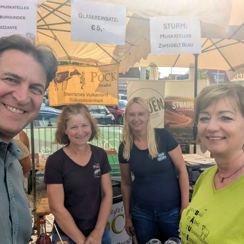 Three women and a man are standing under a tent with signs advertising drinks and food. The woman on the right wears a green shirt, and the others wear black. They are smiling for a photo.